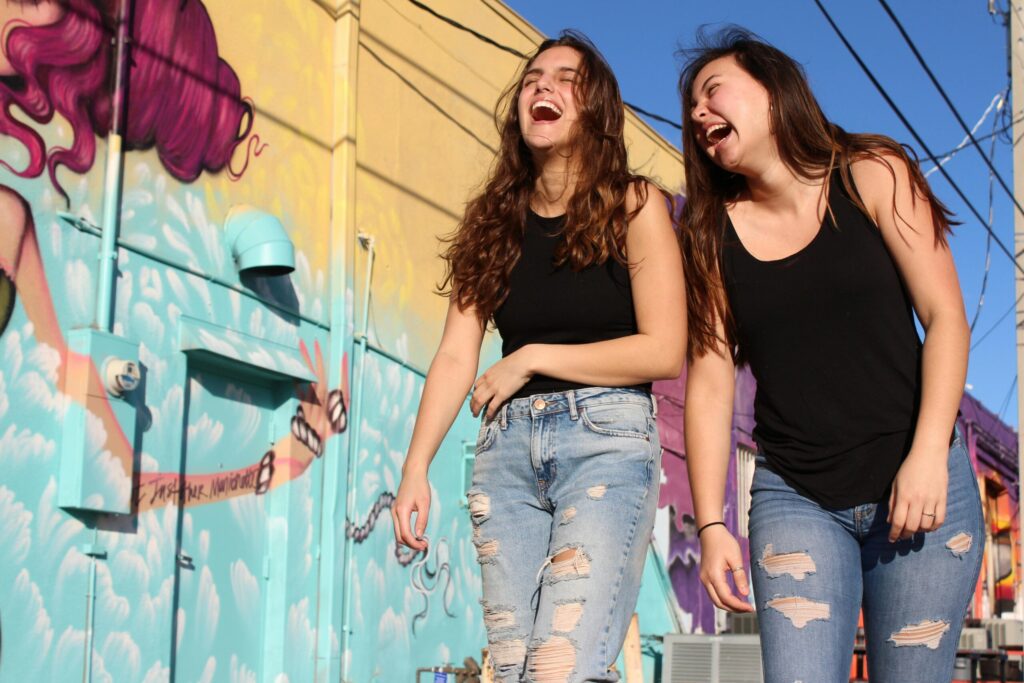 Two young women laughing together outdoors beside a colorful mural wall, enjoying recovery and friendship in Asbury Park, NJ.