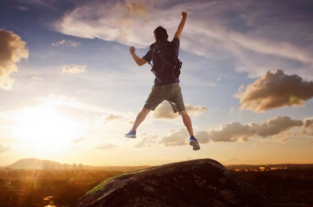 Man with backpack jumping triumphantly on a rocky hilltop against a sunset sky, symbolizing freedom and recovery.