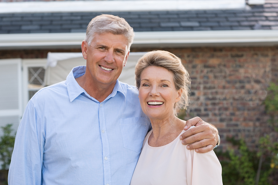 Smiling mature couple standing together outdoors in front of a brick home, embracing and looking hopeful.