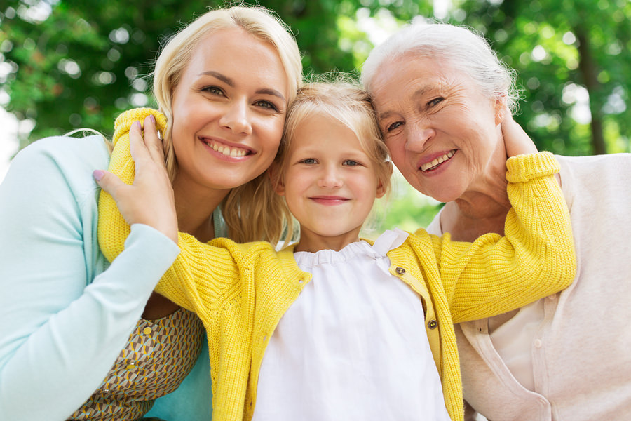 Three generations of women smiling together outdoors, a mother, young daughter, and grandmother embracing.