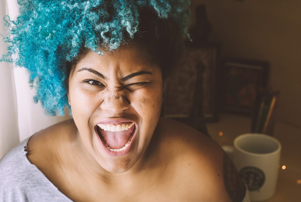 Smiling woman with curly blue hair laughing joyfully in a warmly lit room with a coffee mug nearby.
