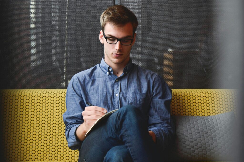 Young man in glasses and blue shirt writing in a journal while seated on a yellow couch during a counseling session.